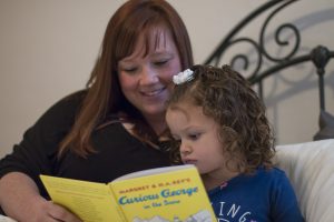 A woman with red hair sits on a bed with a young daughter. They are reading the book "Curious George" together.